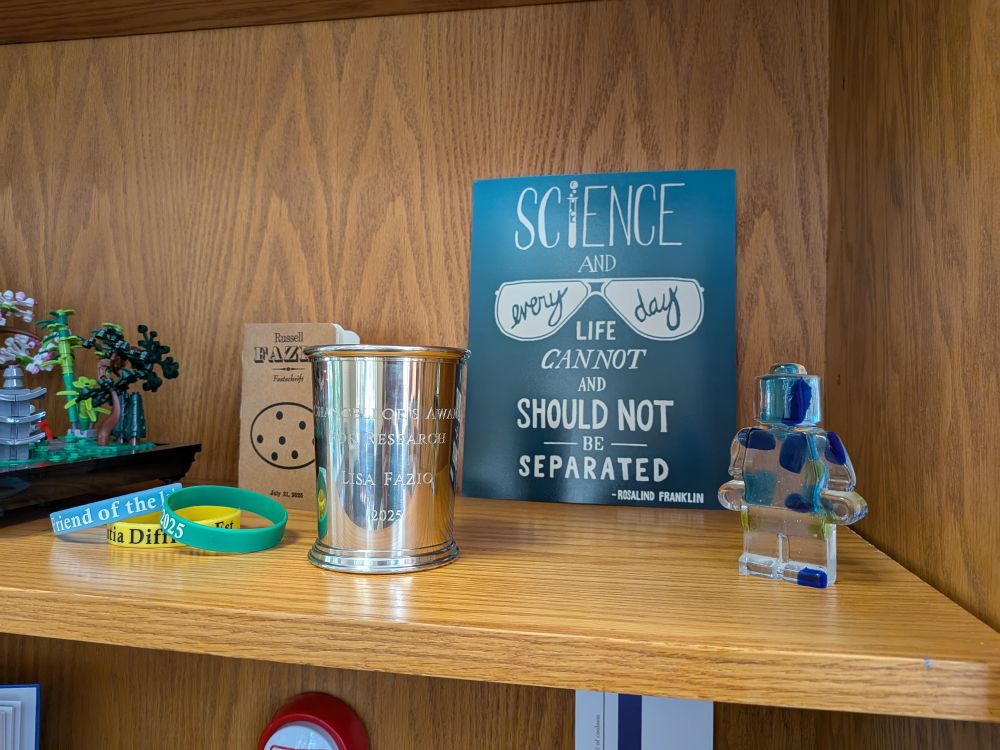 A fancy silver mint julip cup engraved with Chancellor's Award for Research and my name sitting on a wooden shelf with a poster reading "Science and everyday life cannot and should not be separated" - Rosalind Franklin, a glass lego figure, and some other knickknacks 