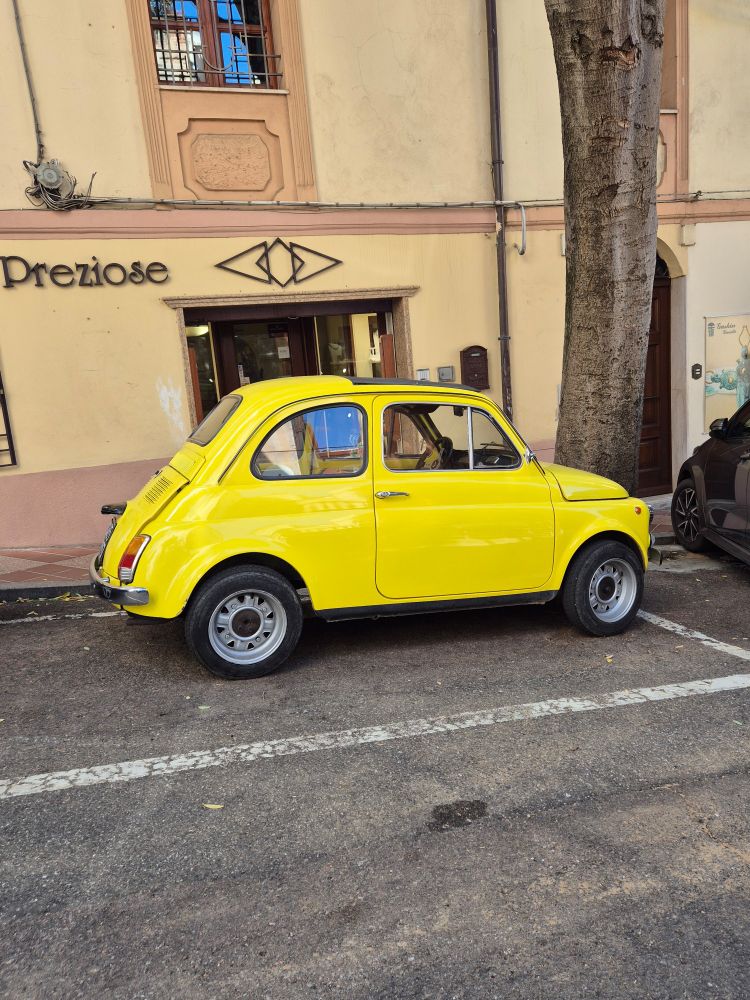 Ein uralter Fiat 500 auf einem viel zu grossen Parkplatz, dahinter ein Gehsteig und eine sardische Häuserfassade, gelbbeigebraun.