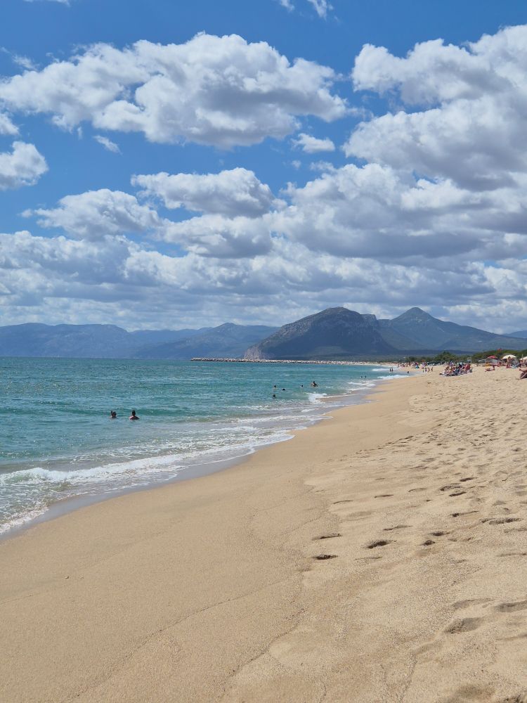Ein langer Sandstrand, Wellen kräuseln sich leicht im Sand, türkisblaugrünes Meer, am Horizont zwei Hügelketten, leicht wolkiger Himmel.