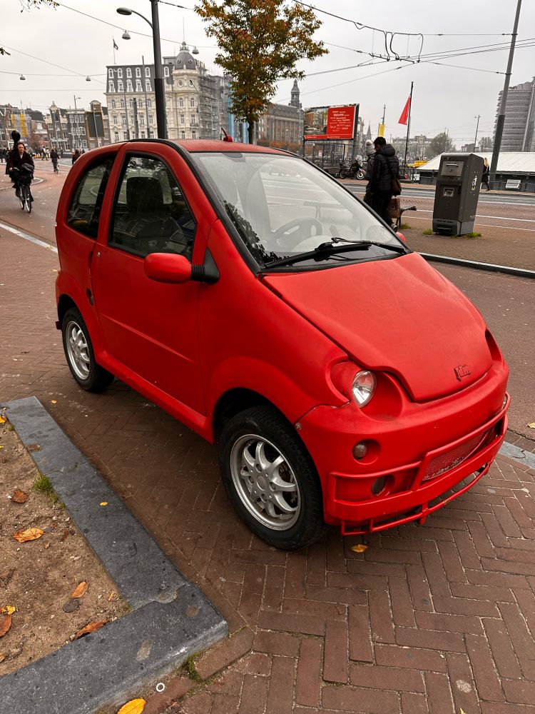 An adorable tiny car - pretty much the size of a wheelchair, parked on a sidewalk but somehow it’s ok. 