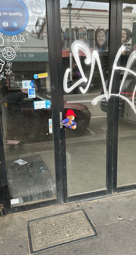 A small bear, maybe Paddington, red hat and pyjamas, stuffed into the door handle of an empty shop.