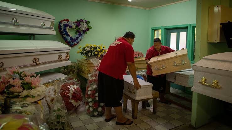 Two men in a room full of funeral requirements, moving very small coffins.