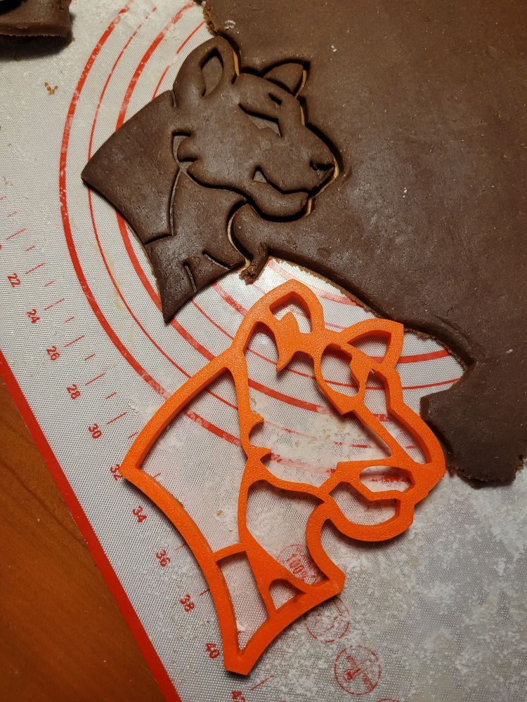 An alternative view of an orange plastic 3D printed gingerbread cutter in the shape of a lioness's head next to a gingerbread cookie cut with that very cutter.