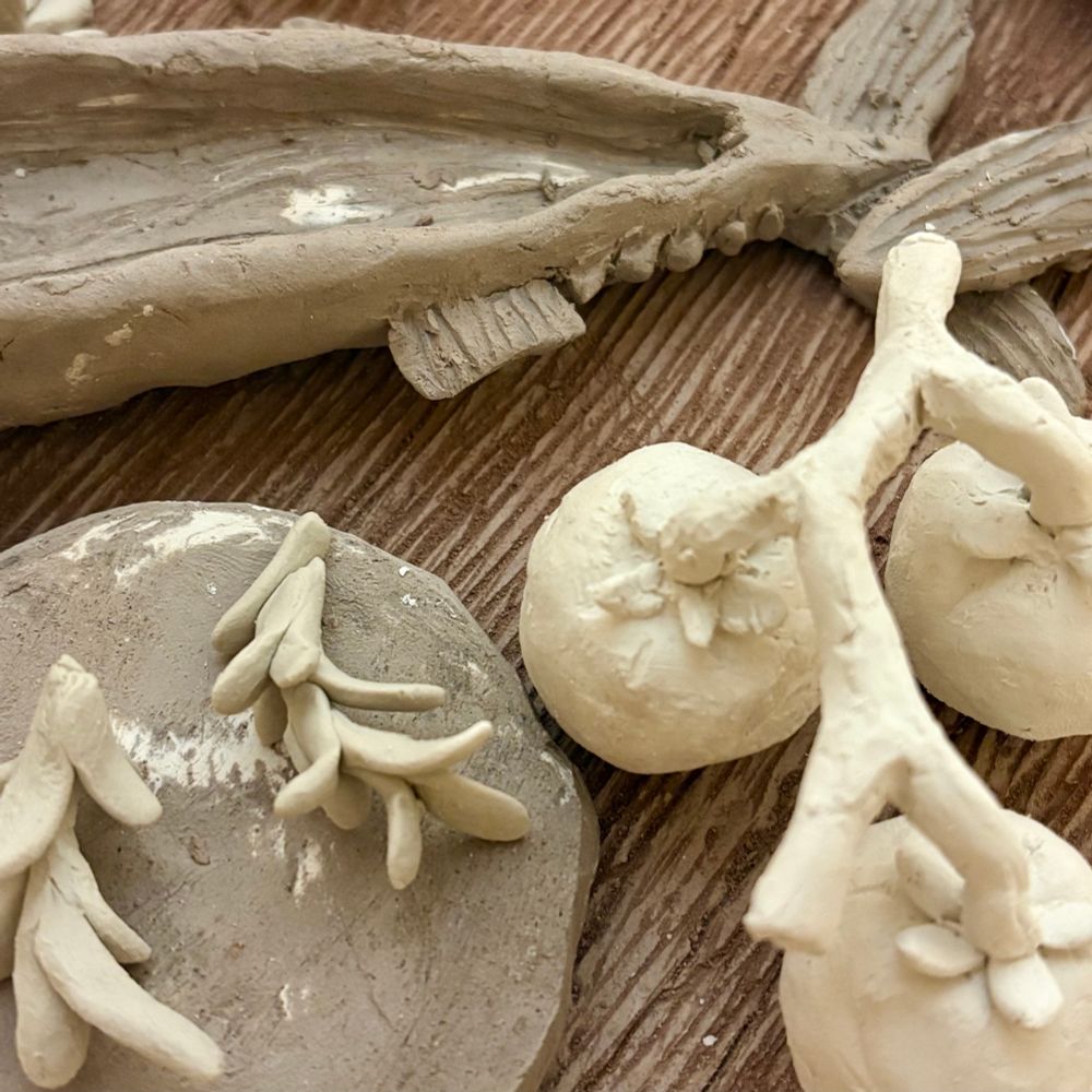 A close up of clay fish sitting in the bottom of a pottery kilns on a red clay cutting board, the fish’s tail is featured, the fish is surrounded by sprigs of rosemary, tomatoes, and clay disks