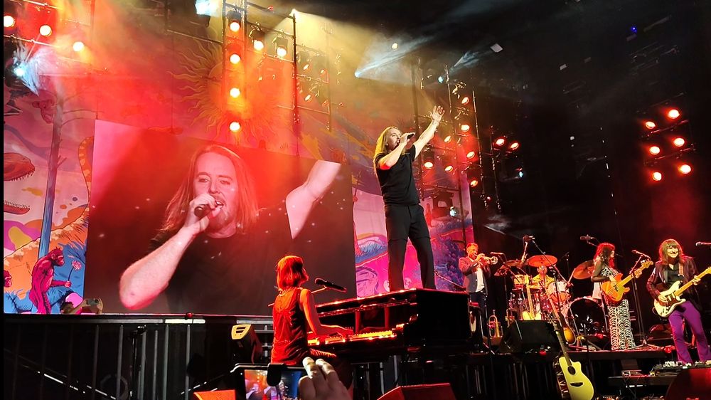 Tim Minchin stood on a piano singing during a finale, with his band playing keys, guitar, bass, trumpet and drums