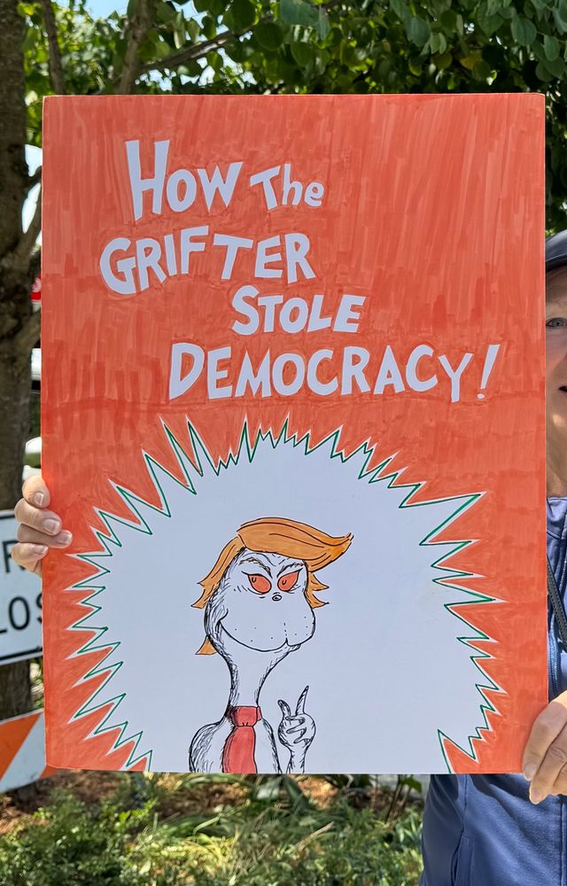 A poster modeled after How the Grinch Stole Christmas with the title changed to How the Grifter Stole Democracy. It has an orange background with white lettering and the Grinch illustration has blonde sweeping hair and a big red tie a la Trump.