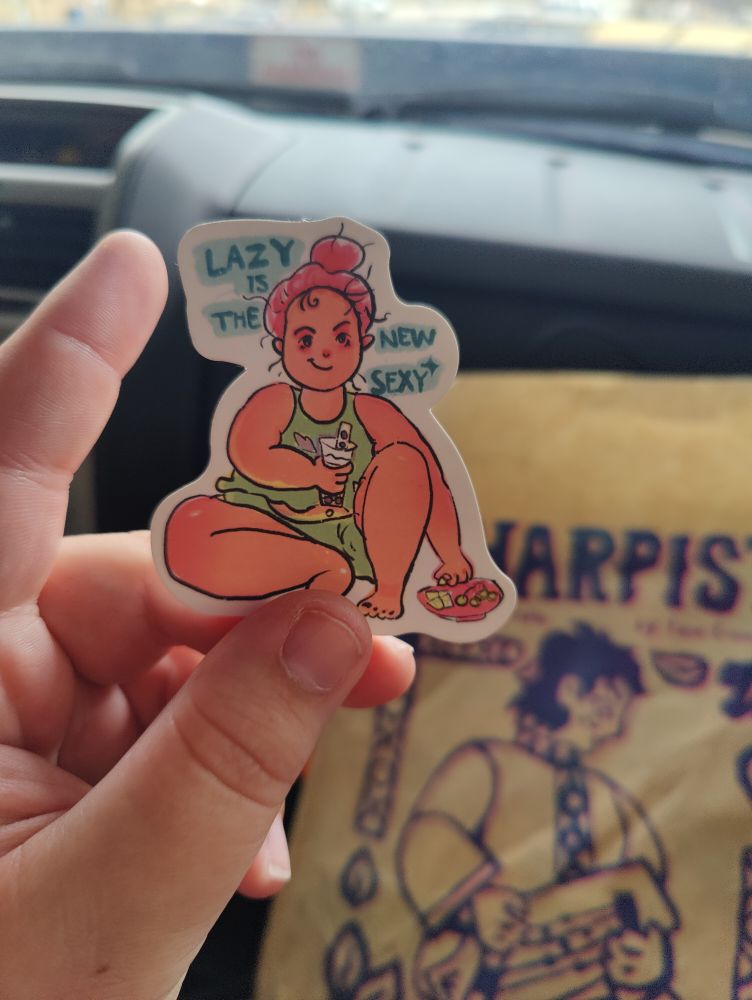 :A sticker of a fat person in a green tank top and green shorts. The person is white with red messy hair and is drinking boba while eating off of a small charcuterie board. The sticker reads: "Lazy is the new sexy"