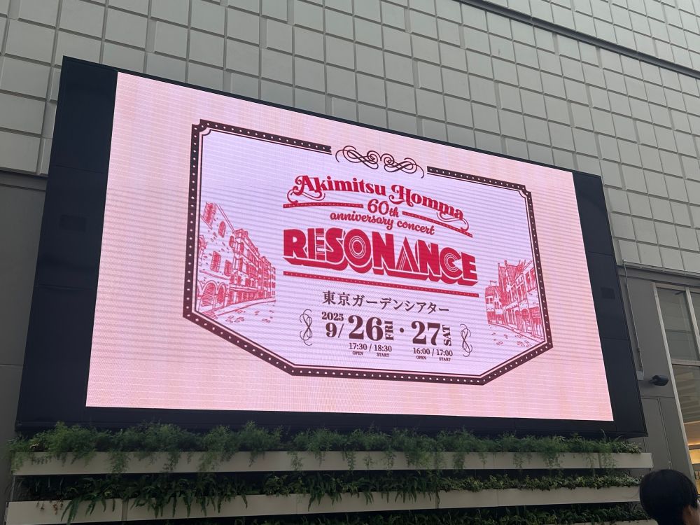 屋外。外は明るい。特大モニターに「Akimitsu Homma 60th anniversary concert “RESONANCE”」のロゴが映し出されている。