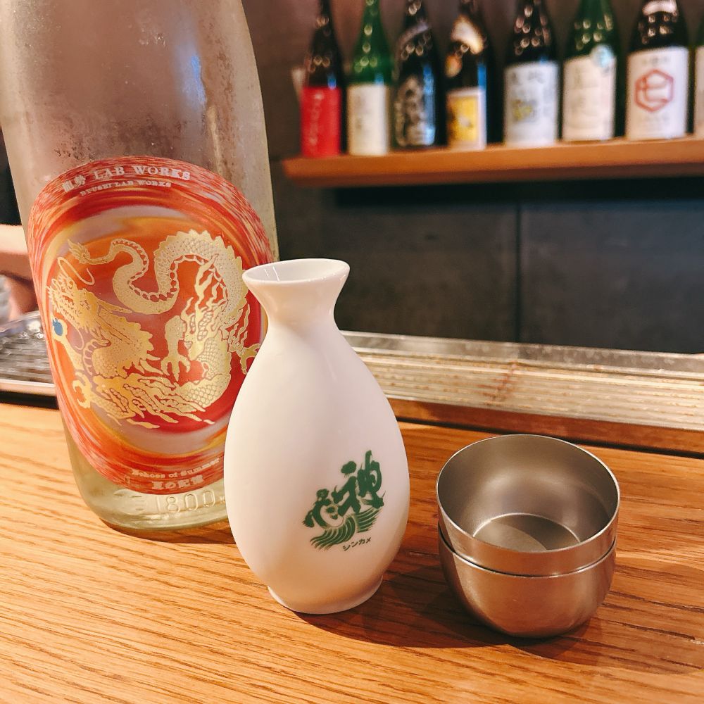 日本酒の一升瓶ととっくり、お猪口が二つ机に置かれている。日本酒のラベルは赤く渦巻く背景の中心に金色の龍が猛々しく浮かぶデザインになっている。金色の部分は多分、箔。