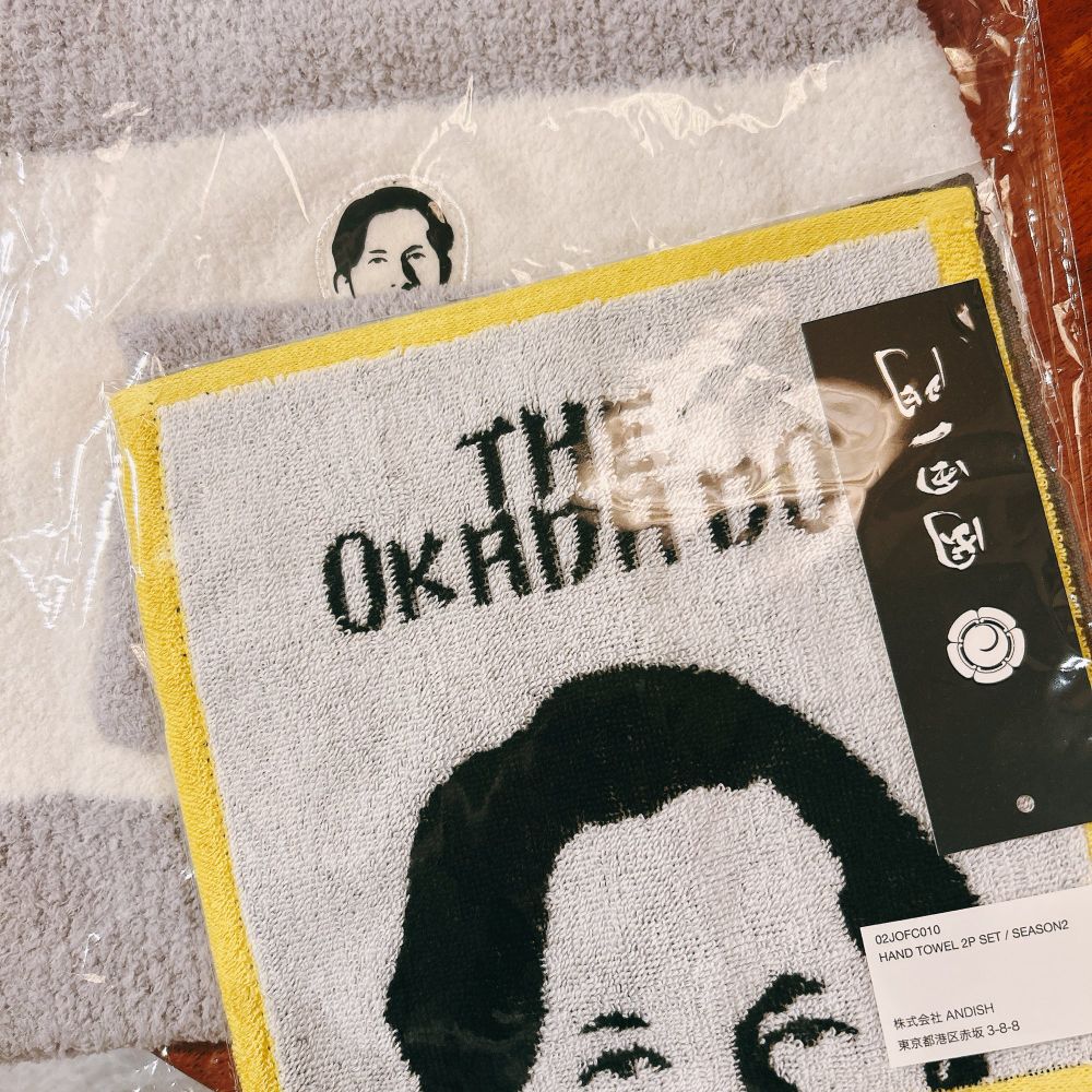 「岡田一門」のタグがついたタオルハンカチ。顔半分をひょっこりと出した、岡田師範のイラストが描かれ、その上に「THE OKADA DO」という文字が書かれている。タオルハンカチの下には腹巻きがある。お腹のポケット部分から、岡田師範のイラストがひょっこりと顔を出している。