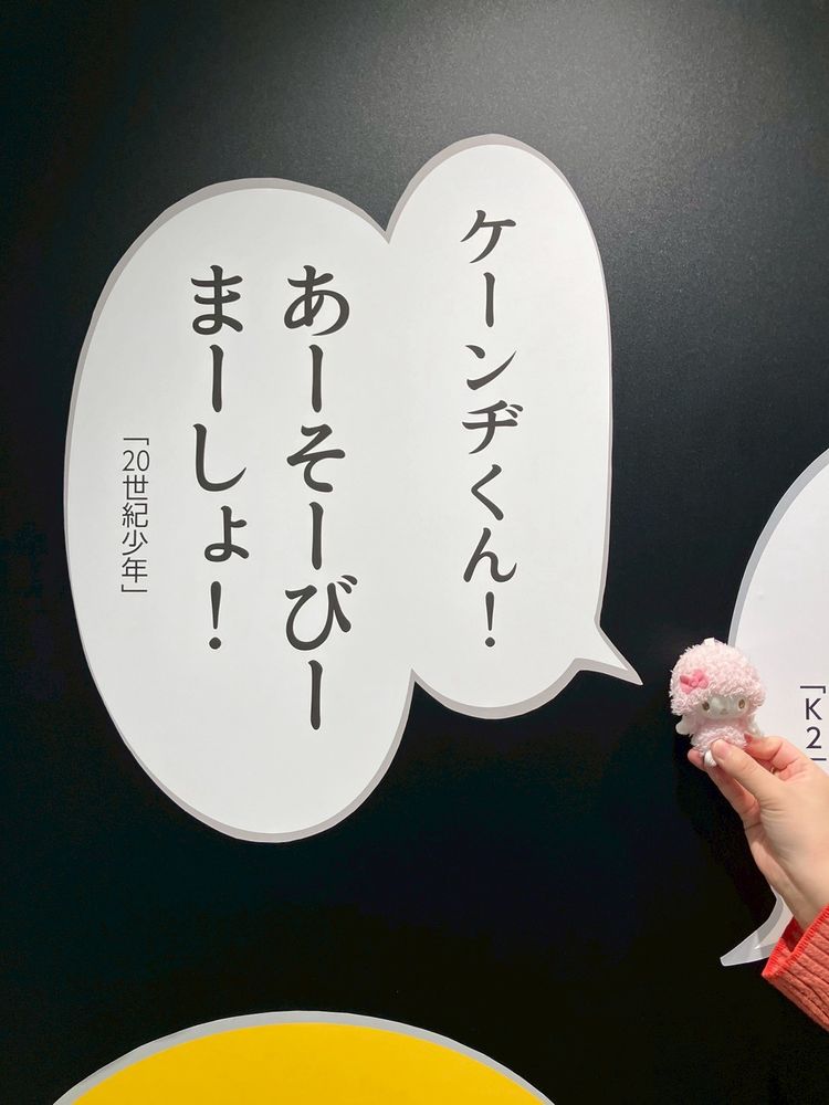 黒い壁に書かれたセリフの吹き出しの前にピアノ先生のぬいぐるみが置かれている。セリフには「ケーンヂくん！あーそーびーまーしょ！「20世紀少年」」と書かれている。