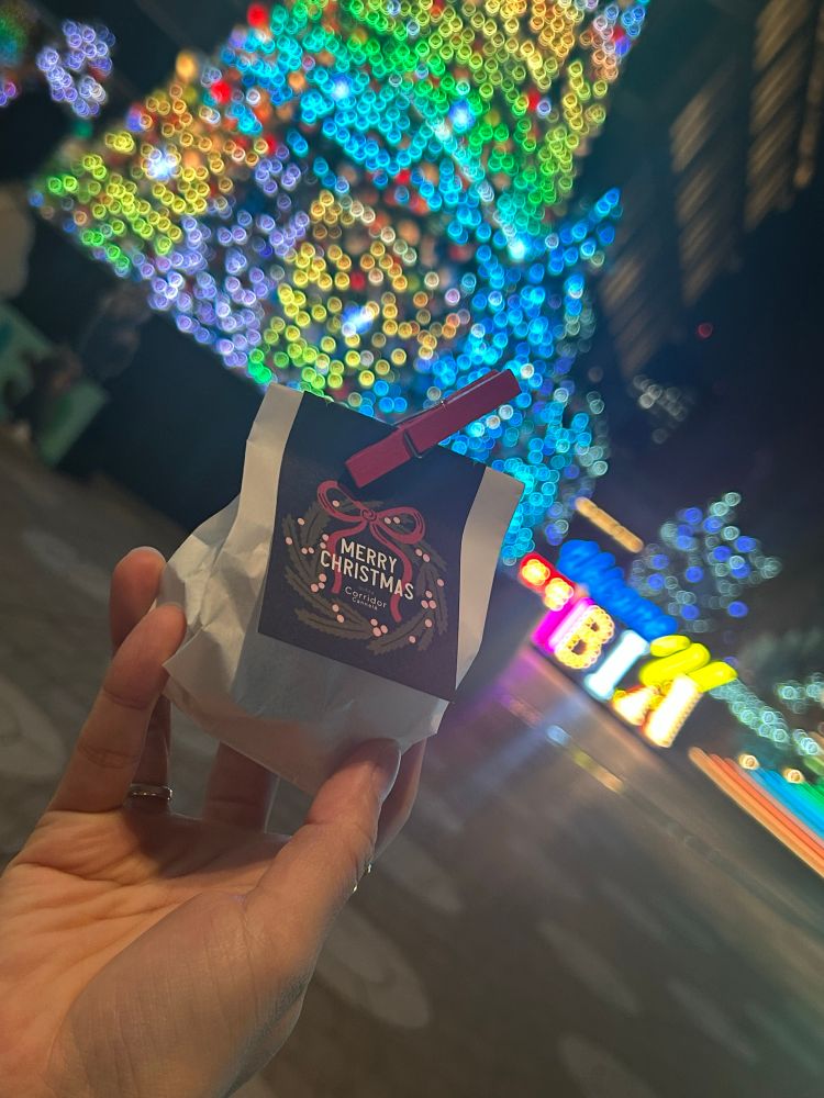 クリスマスツリーのライトアップを背にして、手のひらサイズの袋を見せつける手。袋にはクリスマスリースの絵と「MERRY CHRISTMAS」と書かれた紙が赤い木のクリップで挟まれている。