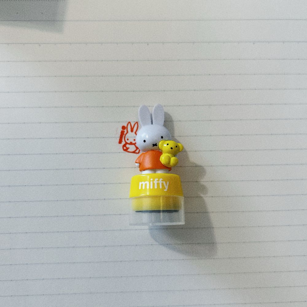 ミッフィーのスタンプ。ミッフィーが黄色いクマのぬいぐるみを抱いて立っているオブジェがついている。後ろのノートにハンコが一つ押されている。