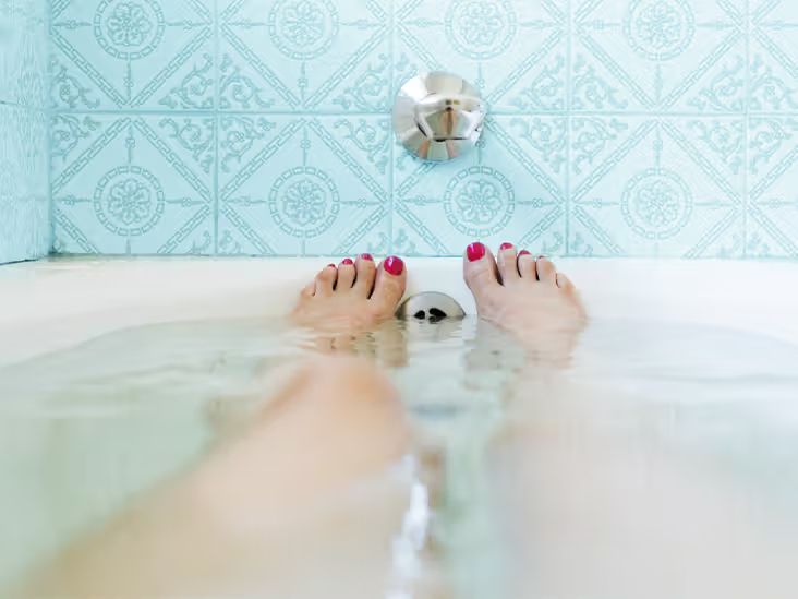 An image of a woman's legs stretching out towards the end of a full bathtub. The woman's toenails are painted bright red. There are blue tiles  on the wall above the tub.