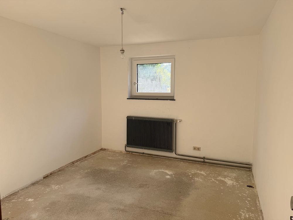 An empty room with white walls, bare concrete floor, a single window, radiator and dangling light bulb 