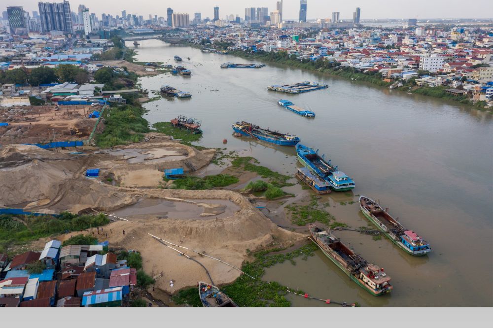 A drone shot shows numerous barges off-loading river sand to the shore of the Bassac River, with Phnom Penh's skyline in the background. Photo credit: Andy Ball