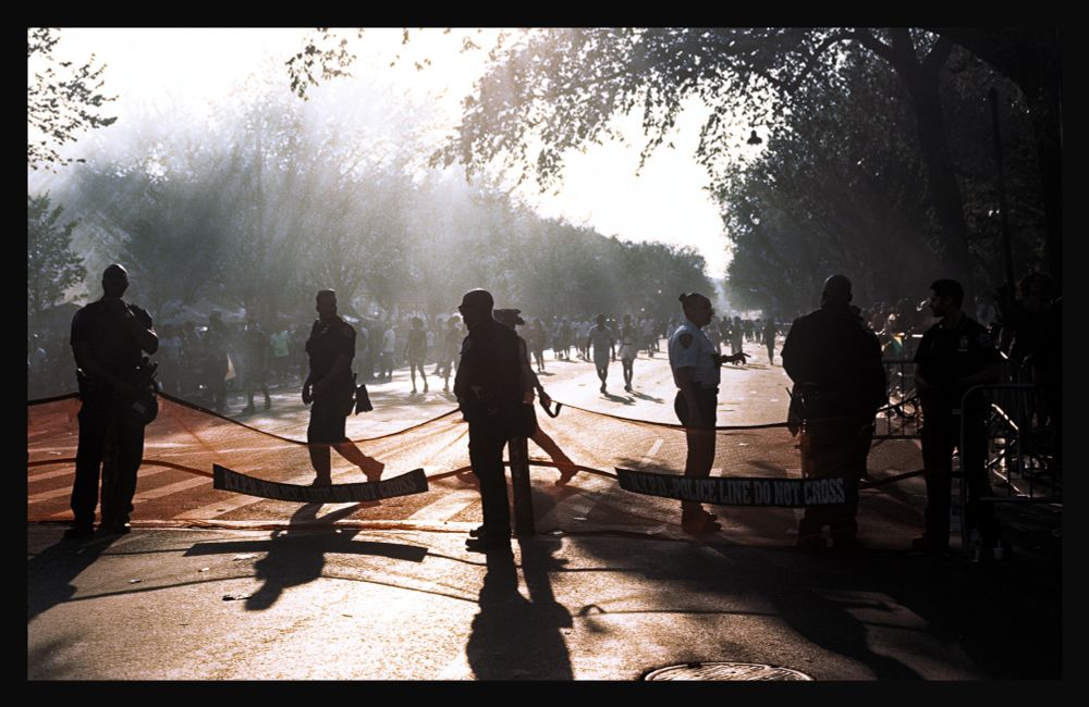 a half-dozen NYPD officers stand on either side of waist-high orange mesh net with "Police Line Do Not Cross" written on it, backlit by afternoon sun mixing with the smoke from grills and cookers. West Indian American Day Parade attendees in the road behind and filtering into congested sidewalks to get by.