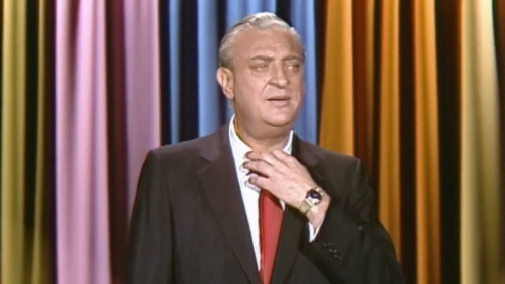Rodney Dangerfield in front of multi-colored curtains, adjusting his collar