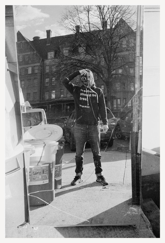 Black and white self-portrait in a mirror sitting on the sidewalk in Brooklyn. Camera held up to my eye in one hand, hand-rolled cigarette in the other. My hair falls past my face to the shoulder of my black hoodie over dark jeans and Nike Air Max Pre-Day sneakers.