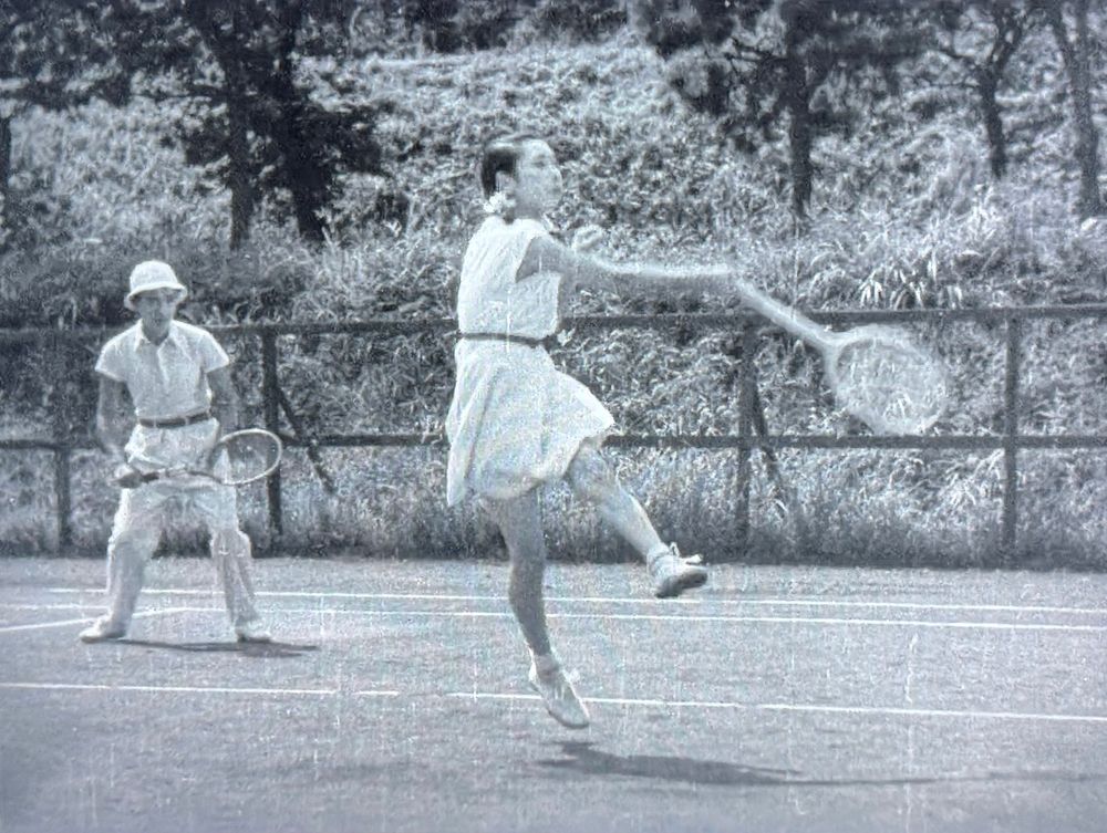 Legendary actress jumping and landing on one foot as follows through slamming the ball in mixed doubles as her lucky partner looks on. B&W 

Phoenix Kinoshita 