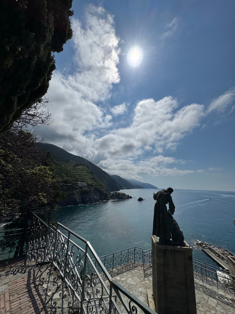Iron wrought stairs w statue leaning down looking over Mediterranean and coast and hills w a sun and clouds. Blue sky & blue green water