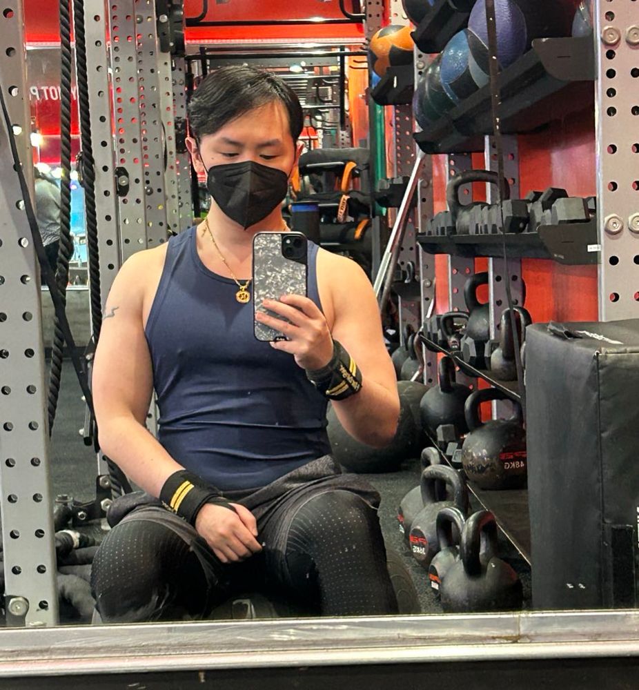 A picture of me sitting on a yoga ball at the gym, wearing a black KN95, a navy tank top, and black shorts with black leggings 