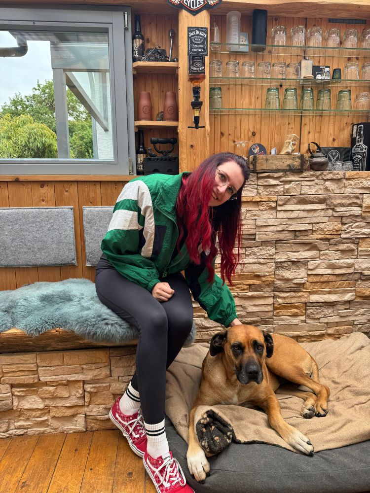 Foto von mir neben einem großen süßen braunen Hund (kenne die Rasse nicht). Sie heißt Miley. Hinter mir stehen ganz viele Gläser 