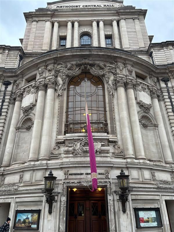 Facade of the Central Hall in London, featuring ornate stonework and a tall purple banner.