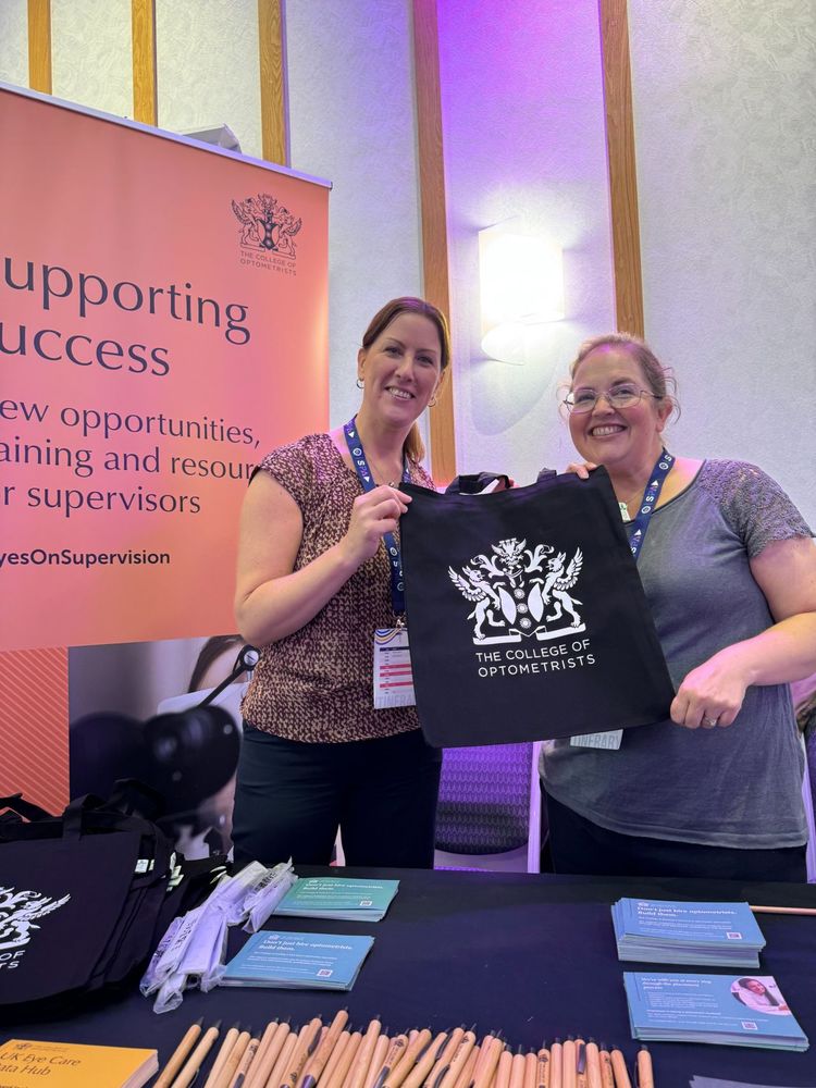 Jo Powel and Sandra Holmes stand at an exhibition booth for The College of Optometrists, smiling and holding a promotional tote bag featuring the college's logo.
