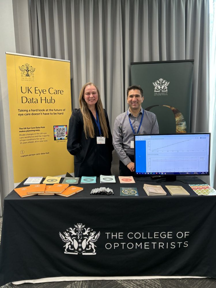 College staff Juliet Leach and Neal Suchak standing in front of a promotional banner for the UK Eye Care Data Hub at a booth hosted by The College of Optometrists, displaying informational materials and a monitor showing a data graph.