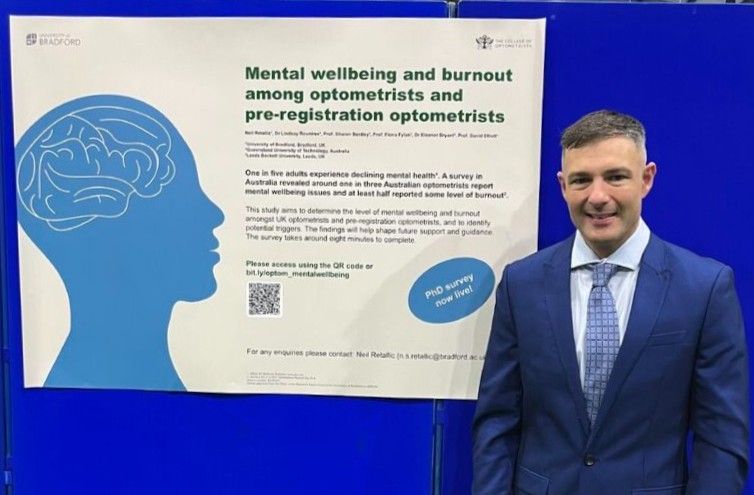 Neil Retallic in a blue suit standing next to a presentation poster about mental wellbeing and burnout among optometrists at a conference.
