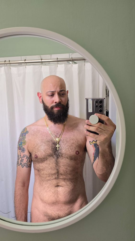 Shirtless, bald, bearded man takes a mirror selfie in a bathroom, wearing gold necklaces, with colorful tattoos on both upper arms. A white shower curtain and round mirror frame are visible in the background.