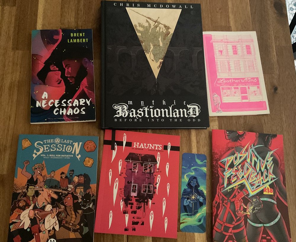 Books, zines, and an illustrated bookmark displayed on a table:
A Necessary Chaos by Brent Lambert
Mythic Bastionland by Chris McDowall
Leatherbound by Ben Mehlos
The Last Session Volume 1 by Walls, Dozerdraws, and Myers
Haunts by Kaylee Rowena
Positive Feedback by Ben Mehlos
