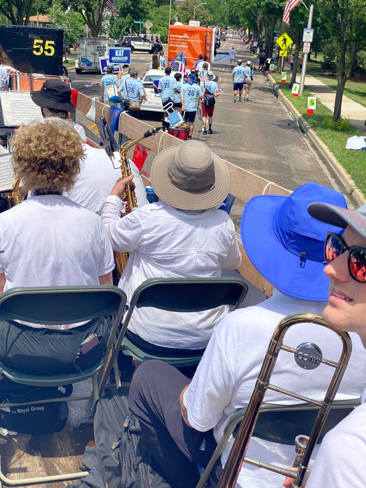 A view of the Kat car and volunteers from the trombone section of the parade entry immediately following it. Shows the parade route, a dozen volunteers walking, dressed in blue, carrying signs. 