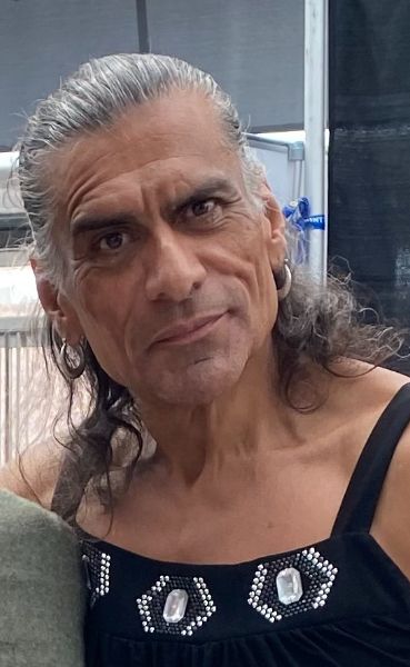 A photo of Johnathan Joss from a con, with long, wavy grey hair. He is wearing a black tank with rhinestone accents.