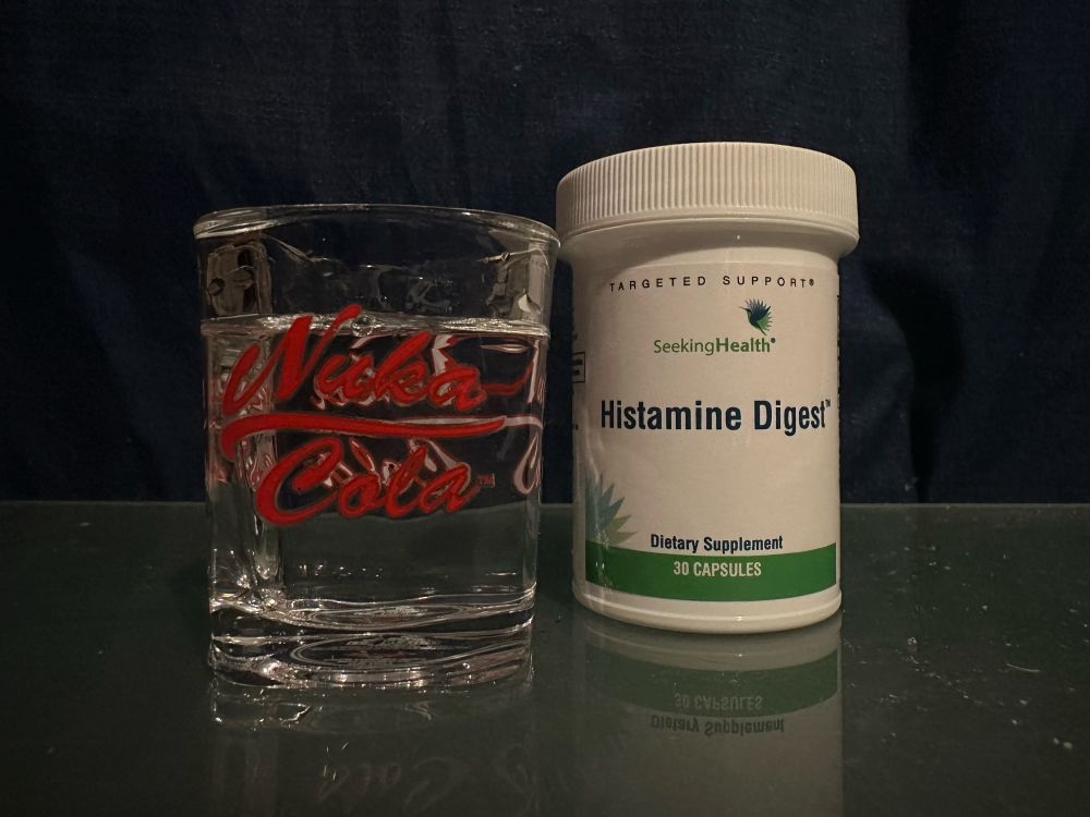 A photo of a shot glass that says “Nuka-Cola” filled with a clear liquid next to a supplement bottle that reads “Histamine Digest”