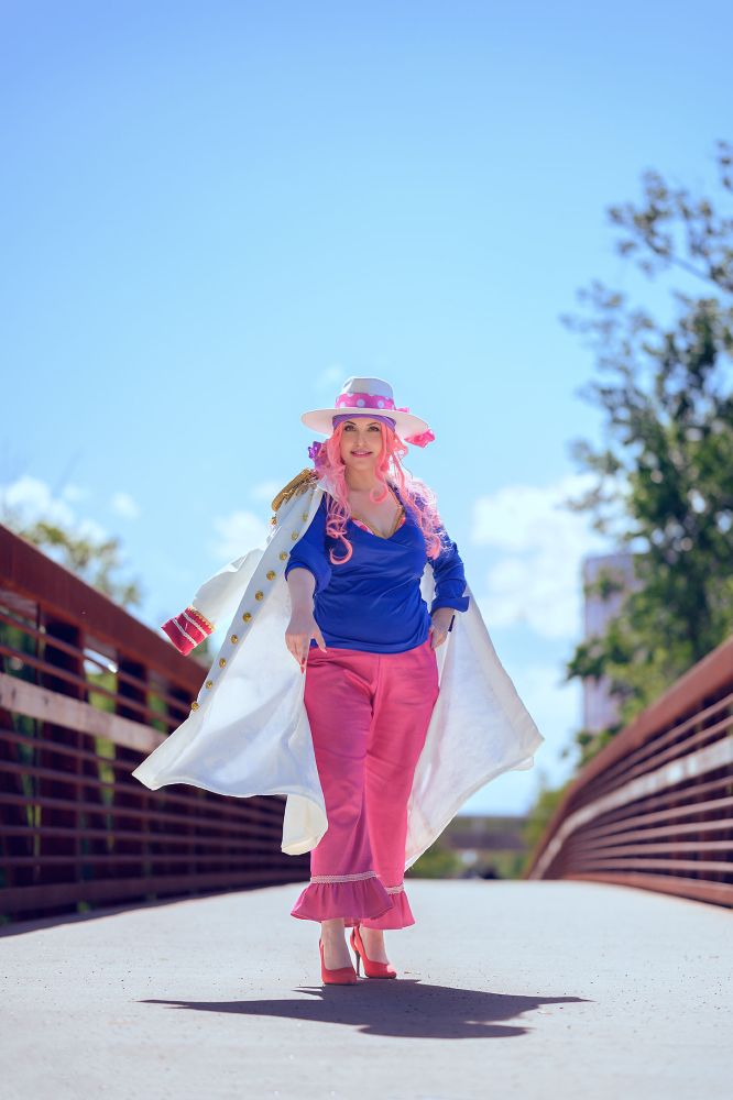 A person in a bright outfit with pink hair walks confidently on a bridge. She wears a blue top, pink pants, and high heels, complemented by a wide-brimmed hat and a long white coat. The scene features a clear blue sky and green trees in the background.