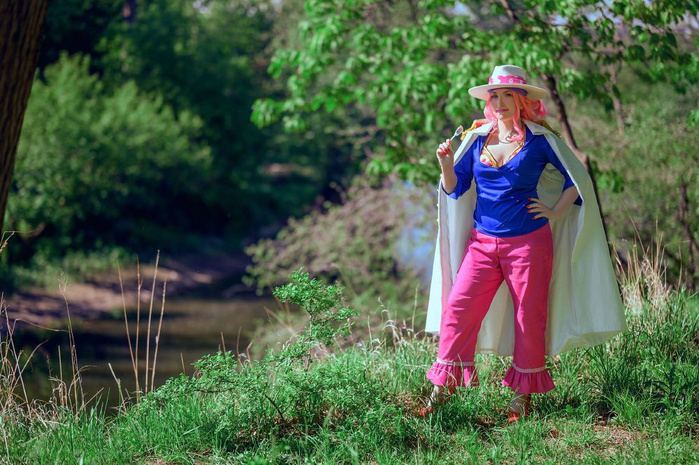 A person stands confidently by a riverbank, dressed in a blue top and pink pants with ruffled hems. She has long pink hair complemented by a wide-brimmed hat and a long white coat. She is holding a lollipop. Lush green trees and grass surround her, creating a natural outdoor setting.