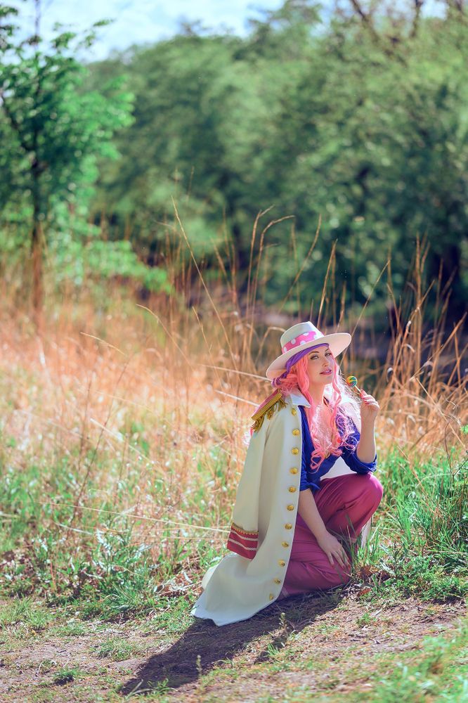 A person with pink hair is kneeling in a grassy area, wearing a wide-brimmed hat and a white coat with red accents. They are dressed as a young Big Mom from "One Piece," surrounded by tall grass and greenery. They are smiling and holding a lollypop.
