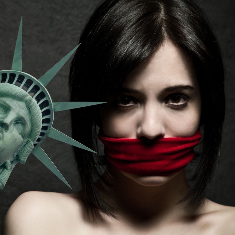 Image is of a woman with dark hair in a straight bob. She looks directly at you. She has dark eyes and is wearing a red cloth gag over her mouth. The head and crown of the statue of liberty is poking in from the side.