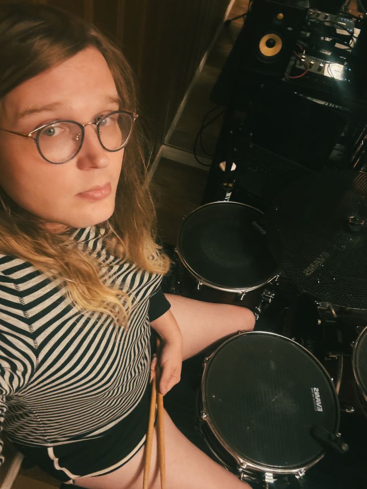 a tall, slightly heavyset blonde girl behind a drum set. she’s wearing a gray and white striped  long sleeve shirt and black and white dolphin shorts and looks relaxed 