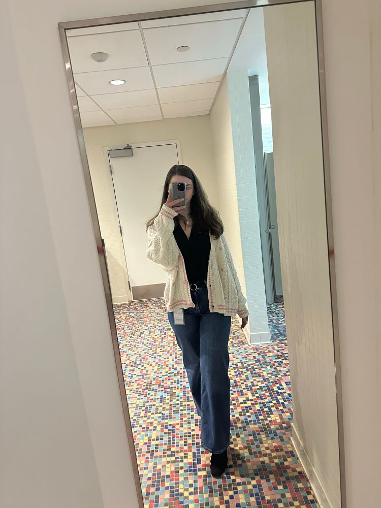 a full body photo of a white woman with brown hair wearing dark denim wide leg jeans, black boots, a black blouse and the Taylor Swift Lover cardigan 