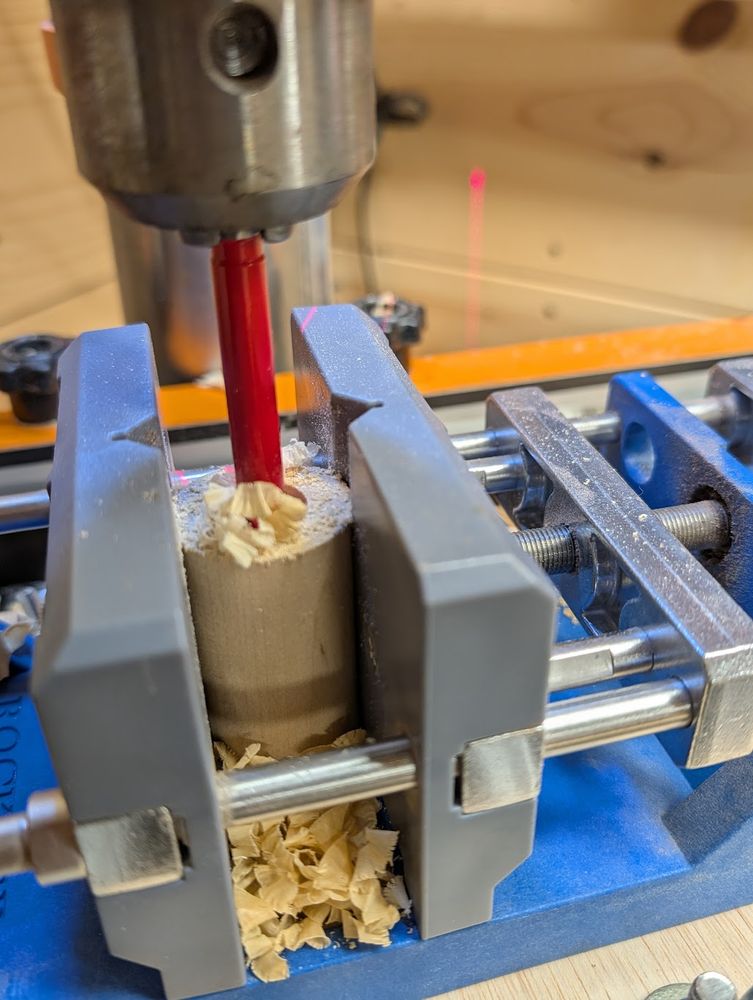 A piece of birch wood mounted in a drill press, being bored out to prepare for the installation of mounting hardware.