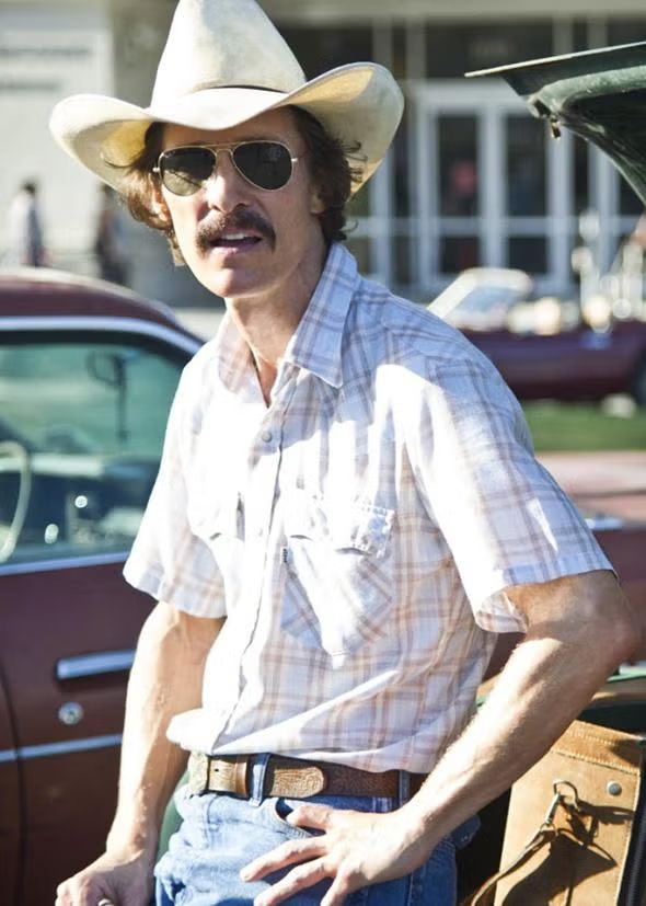 Image Matthew McConaughey as Ron Woodroof in the Dallas Buyers Club. Matthew McConaughey is wearing a straw cowboy hat, sun glasses, long 70's hair, mustache, white button down short sleeve shirt. He looks very sickly, skinny, pale, flat chested, and generally un healthy.   