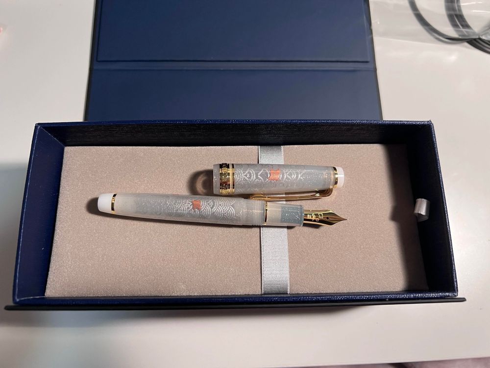 Photo of a transparent fountain pen with Japanese sea designs and silver sparkles. Both on the cap and the body you can see a small coppery stingray. The nib and ring are gold, finials are white 