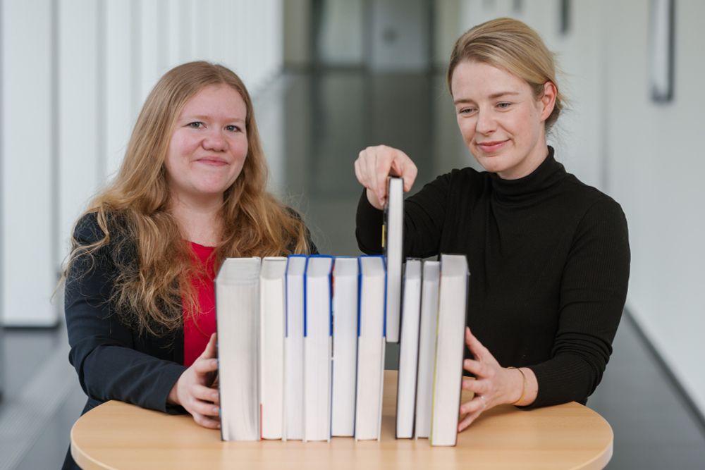 Zwei Forscherinnen stehen hinter einer Bücherreihe und ziehen ein Buch heraus.