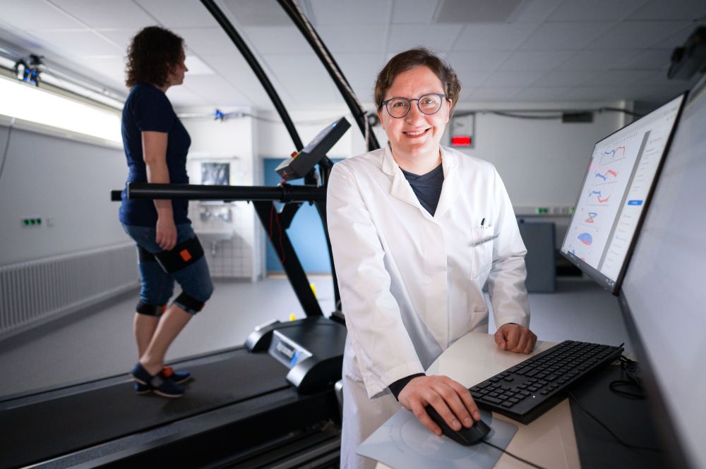 Professorin Bergita Ganse steht an einem Bildschirm, im Hintergrund ist eine Versuchsperson auf dem Laufband.