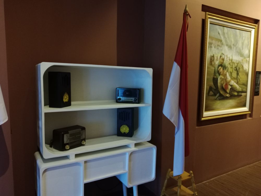 Four old radios in an open white cabinet, continuously broadcasting recordings of the declaration of independence and news from the same year. Next to the cabinet is an Indonesian flag. 