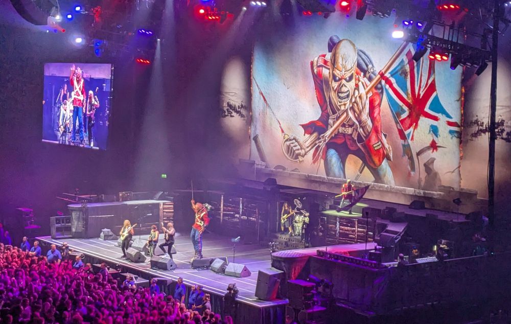 British rock band Iron Maiden on stage. Mascot Eddie behind them, both on stage and on stage, in full "The Trooper" rig - 18th C. British soldier uniform, red tunic, white cross-straps, sword in one hand, British flag in the other.