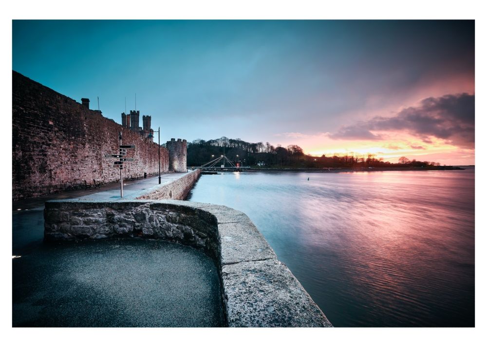 Llun o ran o waliau'r Dre o'r prom yng Ngahernarfon wrth i'r haul fachlud. 
A picture of the town walls in Caernarfon taken on the prom at sunset looking over the Aber bridge